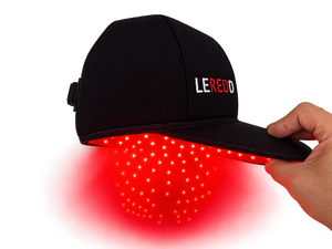 A black LED light therapy cap with the brand name 'LEREDD' embroidered on the side, held by a hand to demonstrate the red light therapy glow emanating from the underside.